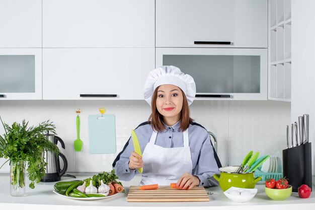 Sonriente mujer chef y verduras frescas con equipo de cocina y picar zanahoria en la cocina blanca