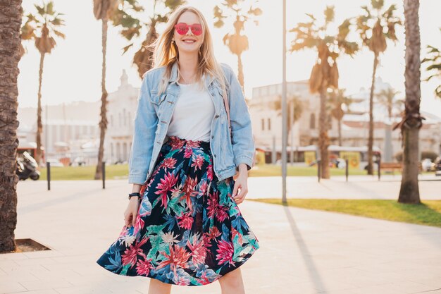 Sonriente mujer coqueteando caminando en las calles de la ciudad con elegante falda estampada y chaqueta vaquera de gran tamaño con gafas de sol rosa