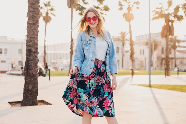 Sonriente mujer coqueteando caminando en las calles de la ciudad con elegante falda estampada y chaqueta vaquera de gran tamaño con gafas de sol rosa
