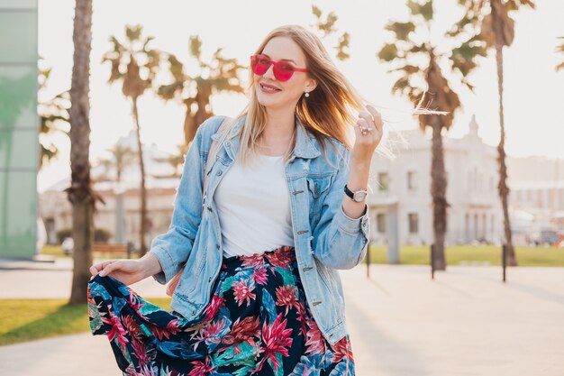 Sonriente mujer coqueteando caminando en las calles de la ciudad con elegante falda estampada y chaqueta vaquera de gran tamaño con gafas de sol rosa