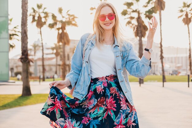 Sonriente mujer coqueteando caminando en las calles de la ciudad con elegante falda estampada y chaqueta vaquera de gran tamaño con gafas de sol rosa