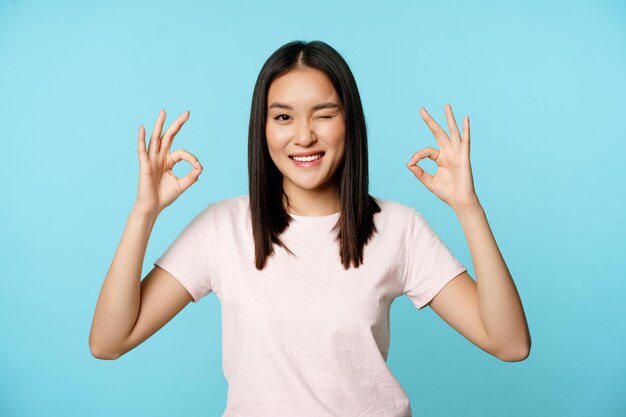 Sonriente mujer coreana guiñando un ojo mostrando signos bien recomendando empresa o tienda de pie en camiseta ove ...