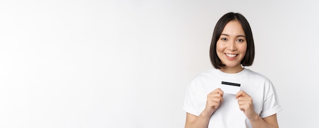 Sonriente mujer coreana mostrando tarjeta de crédito con cara feliz de pie en camiseta sobre fondo blanco.