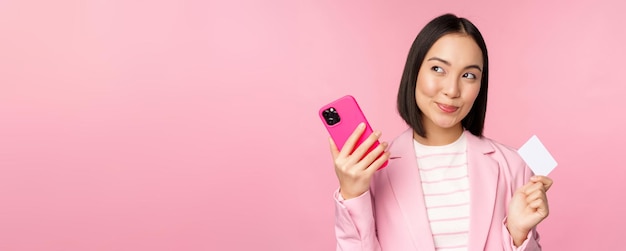 Sonriente mujer corporativa asiática dama en traje pensando en tener un teléfono inteligente y un plan de tarjeta de crédito para comprar algo en línea con fondo rosa de teléfono móvil