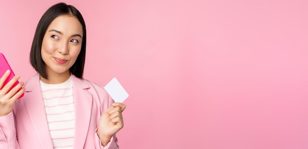 Sonriente mujer corporativa asiática dama en traje pensando en tener un teléfono inteligente y un plan de tarjeta de crédito para comprar s