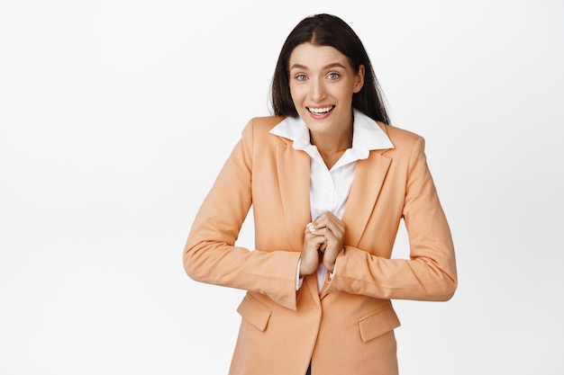 Sonriente mujer corporativa con la esperanza de conseguir algo delicioso luciendo esperanzada de pie en traje contra el fondo blanco.