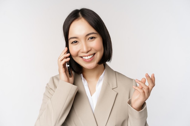 Sonriente mujer corporativa en traje hablando por teléfono móvil con una llamada de negocios en el teléfono inteligente de pie sobre fondo blanco.