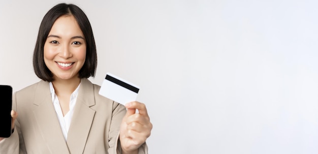 Sonriente mujer corporativa en traje que muestra la pantalla del teléfono móvil y la aplicación en la pantalla del teléfono inteligente de pie sobre fondo blanco