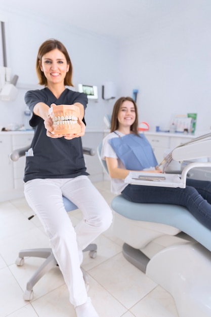 Sonriente mujer dentista mostrando los dientes modelo sentado frente a paciente femenino en la clínica dental