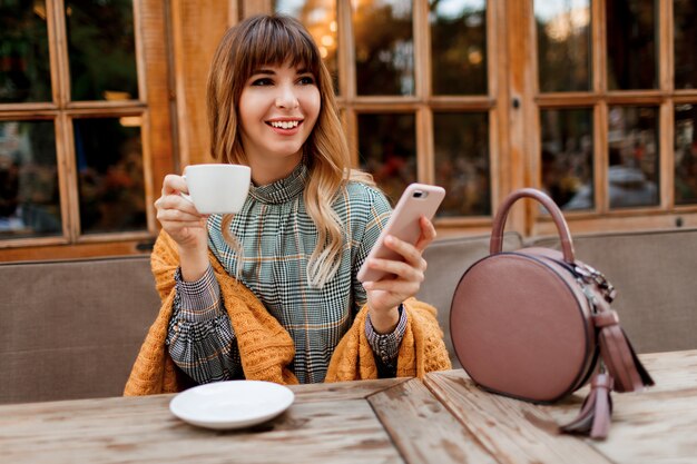 Sonriente mujer despreocupada tomar un café en la acogedora cafetería con interior de madera, mediante teléfono móvil. Sosteniendo una taza de capuchino caliente. Temporada de invierno.