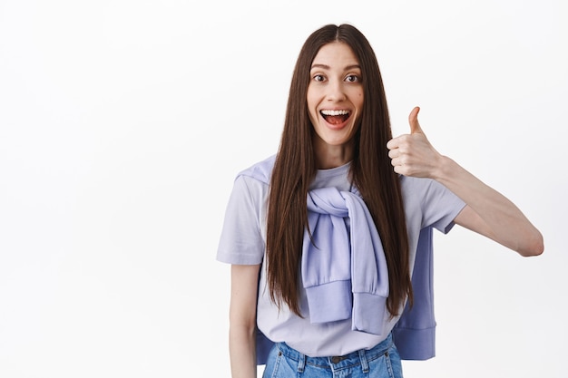 Sonriente mujer divertida con el pelo largo, muestra el pulgar hacia arriba en señal de aprobación, se ve impresionado, elogia el buen trabajo, de pie satisfecho contra la pared blanca