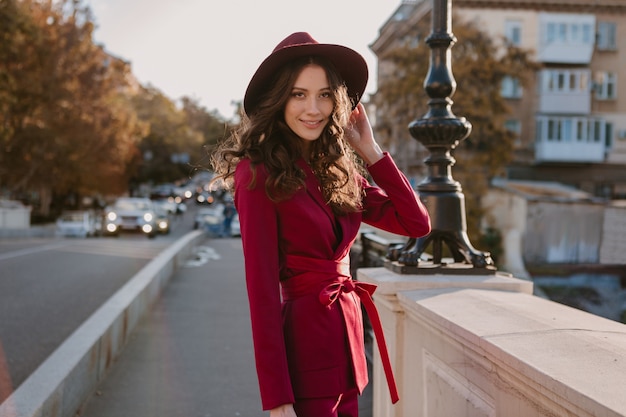 Sonriente mujer elegante hermosa en traje púrpura caminando en las calles de la ciudad, tendencia de moda primavera verano otoño temporada con sombrero, sosteniendo el bolso