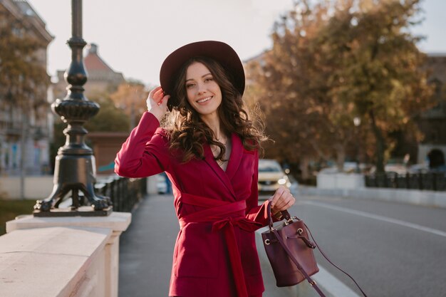 Sonriente mujer elegante hermosa en traje púrpura caminando en las calles de la ciudad, tendencia de moda primavera verano otoño temporada con sombrero, sosteniendo el bolso