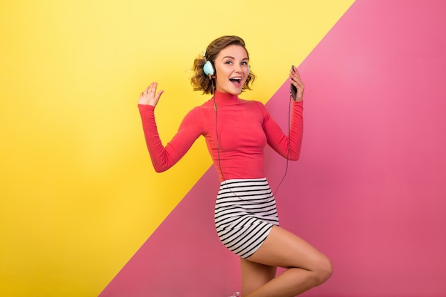 Sonriente mujer emocionada sonriente atractiva en elegante traje colorido bailando y escuchando música en auriculares sobre fondo amarillo rosa, tendencia de moda de verano