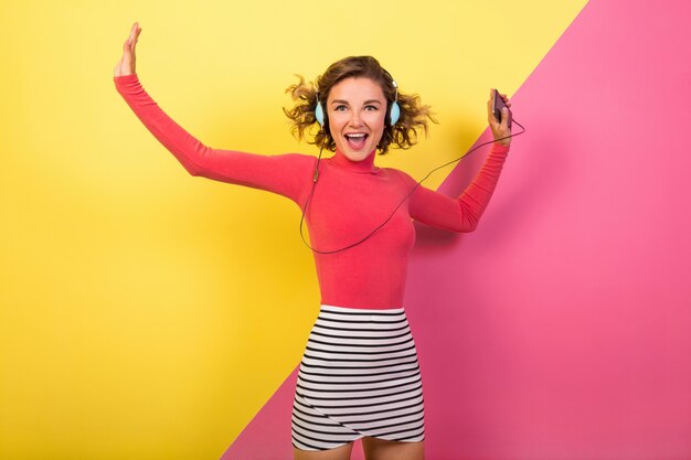 Sonriente mujer emocionada sonriente atractiva en elegante traje colorido bailando y escuchando música en auriculares sobre fondo amarillo rosa, tendencia de moda de verano