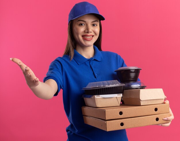 Sonriente mujer de entrega bonita en puntos uniformes con la mano y sostiene el paquete de alimentos y contenedores en cajas de pizza aisladas en la pared rosa con espacio de copia