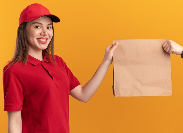 Sonriente mujer de entrega bonita en uniforme da paquete de papel a alguien mirando a la cámara