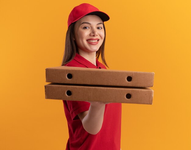 Sonriente mujer de entrega bonita en uniforme se encuentra de lado sosteniendo cajas de pizza en naranja