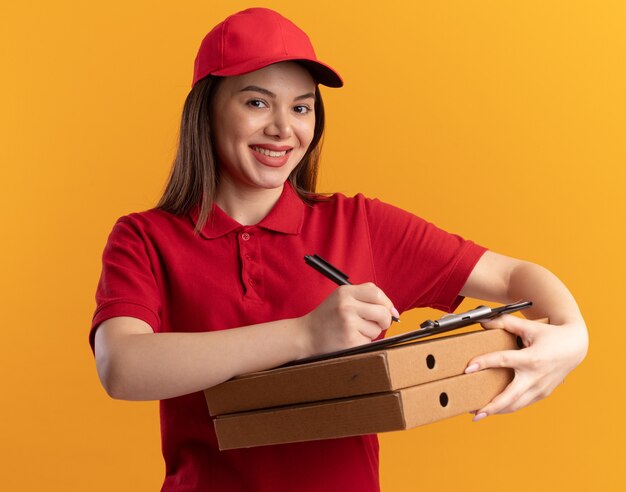 Sonriente mujer de entrega bonita en uniforme escribe en el portapapeles con marcador en cajas de pizza aisladas en la pared naranja con espacio de copia