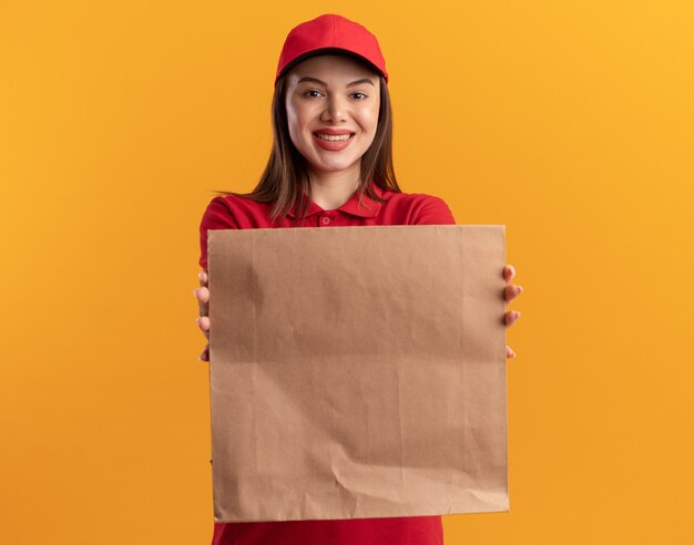 Sonriente mujer de entrega bonita en uniforme estirando paquete de papel en naranja