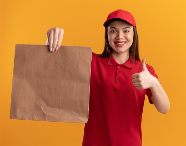 Sonriente mujer de entrega bonita en uniforme hojeando hacia arriba y sosteniendo el paquete de papel en naranja