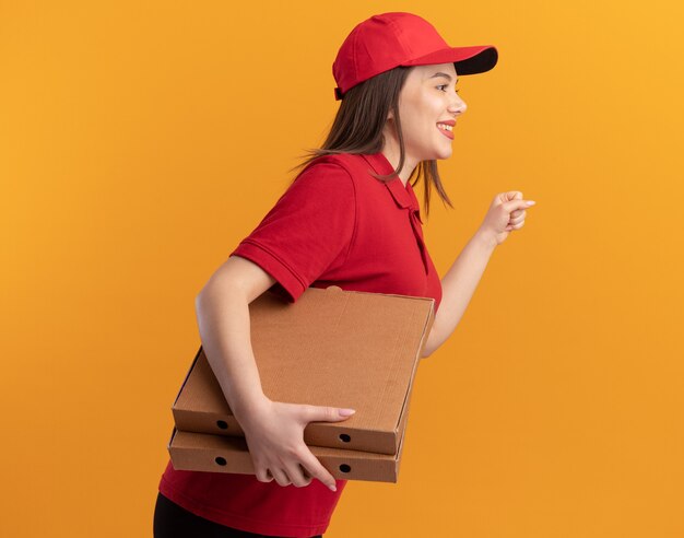 Sonriente mujer de entrega bonita en uniforme se para de lado sosteniendo cajas de pizza y manteniendo el puño aislado en la pared naranja con espacio de copia