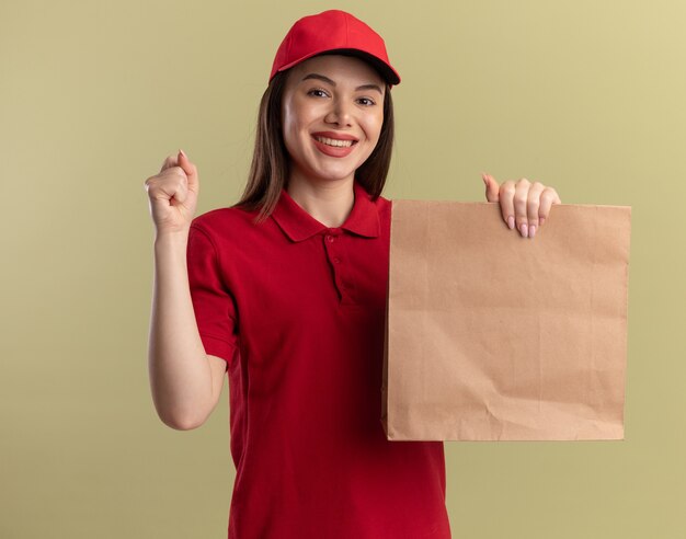 Sonriente mujer de entrega bonita en uniforme mantiene el puño y sostiene el paquete de papel en verde oliva