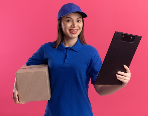 Sonriente mujer de entrega bonita en uniforme sostiene la caja de cartón y el portapapeles aislado en la pared rosa con espacio de copia