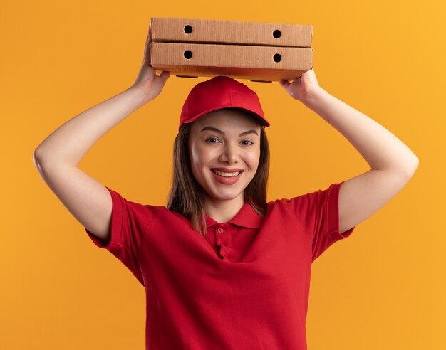 Sonriente mujer de entrega bonita en uniforme sostiene cajas de pizza sobre la cabeza en naranja