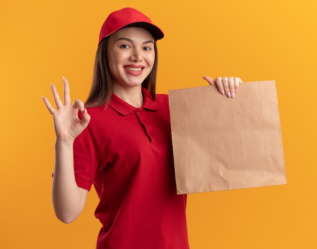 Sonriente mujer de entrega bonita en uniforme sostiene el paquete de papel y gestos ok signo de mano en naranja