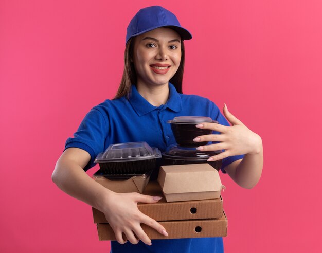 Sonriente mujer de entrega bonita en uniforme sostiene paquetes de alimentos y contenedores en cajas de pizza aisladas en la pared rosa con espacio de copia