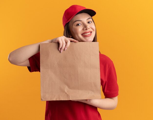 Sonriente mujer de entrega bonita en uniforme tiene paquete de papel aislado en la pared naranja con espacio de copia