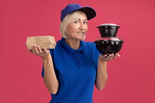 Sonriente mujer de entrega rubia de mediana edad con uniforme azul y gorra sosteniendo paquetes de alimentos de papel y contenedores de alimentos mirando al frente aislado en la pared rosa
