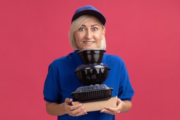 Sonriente mujer de entrega rubia de mediana edad en uniforme azul y gorra sosteniendo paquetes de comida de papel y recipientes de comida poniendo la barbilla sobre ellos