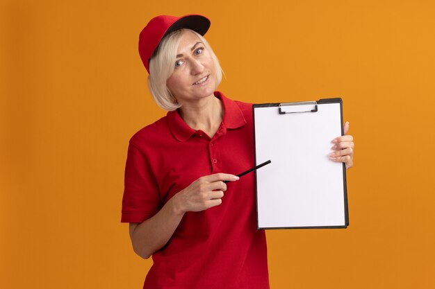 Sonriente mujer de entrega rubia de mediana edad en uniforme rojo y gorra apuntando al portapapeles con lápiz aislado en la pared naranja con espacio de copia