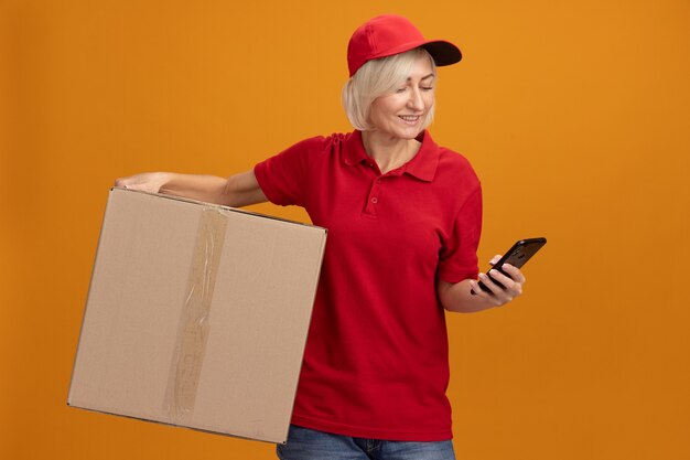 Sonriente mujer de entrega rubia de mediana edad en uniforme rojo y gorra sosteniendo una caja de cartón y un teléfono móvil mirando el teléfono aislado en la pared naranja