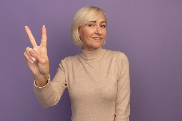 Sonriente mujer eslava bastante rubia gesticulando signo de mano de victoria en púrpura