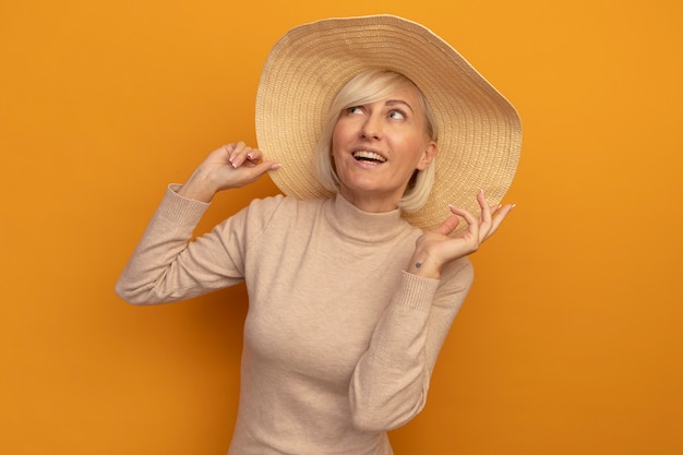 Sonriente mujer eslava rubia bonita con sombrero de playa se encuentra con la mano levantada mirando hacia arriba en naranja