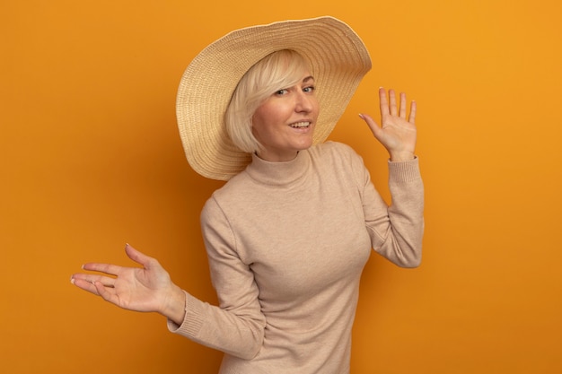 Sonriente mujer eslava rubia bonita con sombrero de playa se encuentra con las manos levantadas en naranja