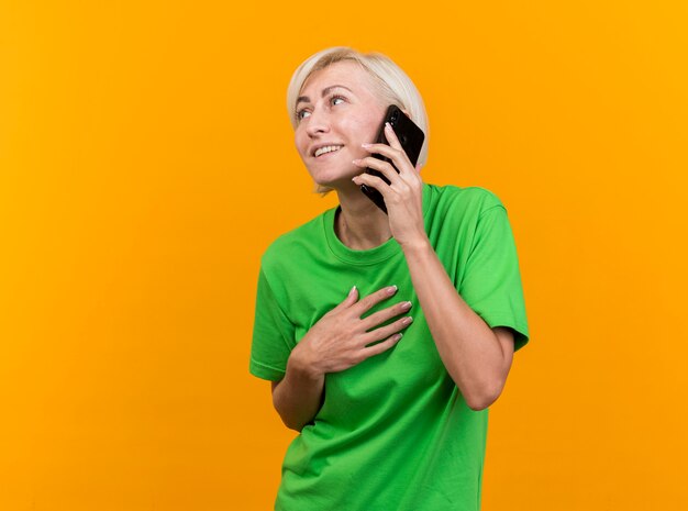 Sonriente mujer eslava rubia de mediana edad hablando por teléfono manteniendo la mano en el pecho mirando al lado aislado sobre fondo amarillo con espacio de copia