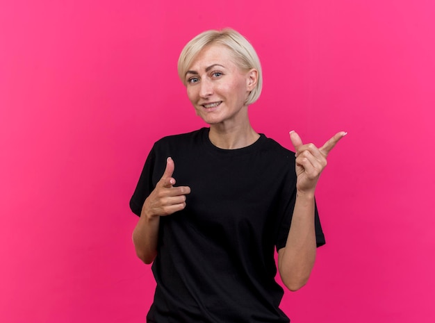 Sonriente mujer eslava rubia de mediana edad mirando al frente apuntando a un lado y a la cámara aislada en la pared rosa con espacio de copia