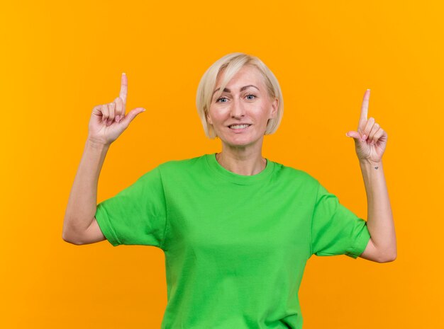 Sonriente mujer eslava rubia de mediana edad mirando a la cámara apuntando hacia arriba aislado sobre fondo amarillo