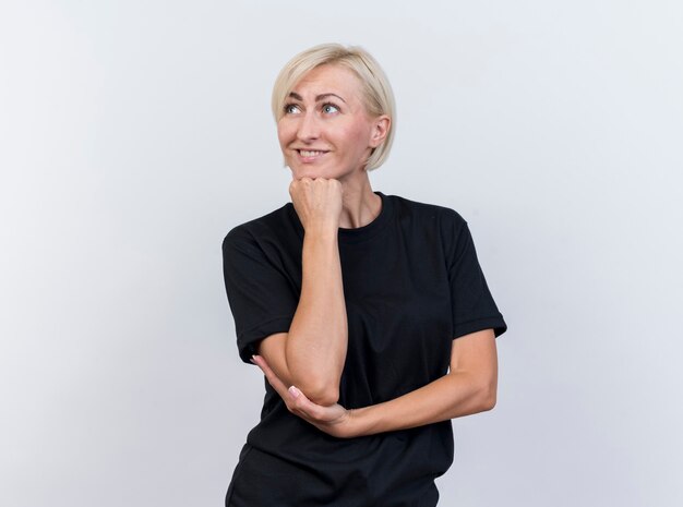 Sonriente mujer eslava rubia de mediana edad poniendo la mano en el codo y en la barbilla mirando al lado aislado sobre fondo blanco con espacio de copia