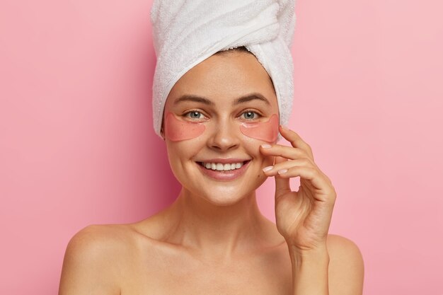 Sonriente mujer europea atractiva con expresión de rostro alegre, usa almohadillas de silicona rosa debajo de los ojos, feliz de verse fresca después de la ducha y los tratamientos de spa, muestra el efecto de una piel perfecta