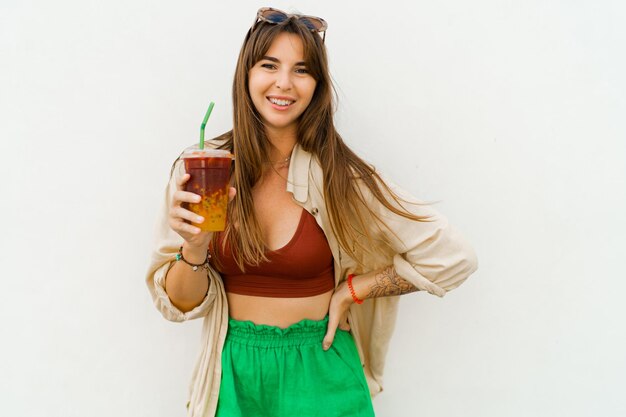 Sonriente mujer europea en elegante traje de verano bebiendo limonada dulce sobre fondo blanco.