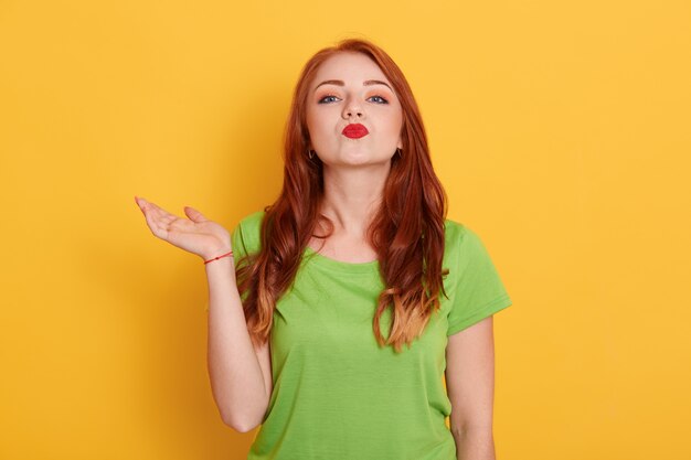 Sonriente mujer europea con encantadora expresión facial vistiendo camiseta verde casual, extendiendo la palma hacia un lado y haciendo gesto de beso