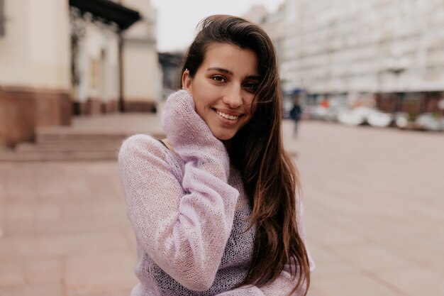 Sonriente mujer feliz con cabello largo vistiendo un suéter violeta posando sobre edificios antiguos