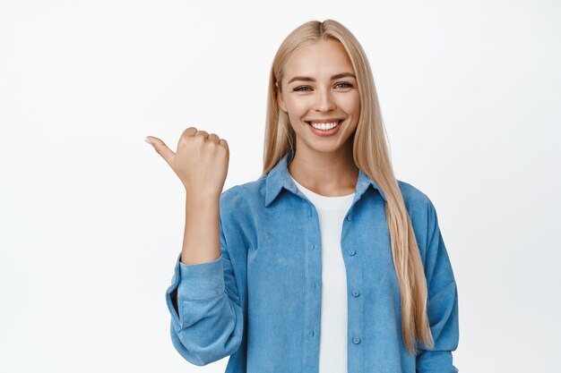 Sonriente mujer feliz con corte de pelo rubio recto, apuntando con el dedo hacia la izquierda y mirando confiado, mostrando el camino, anuncio de venta en blanco.