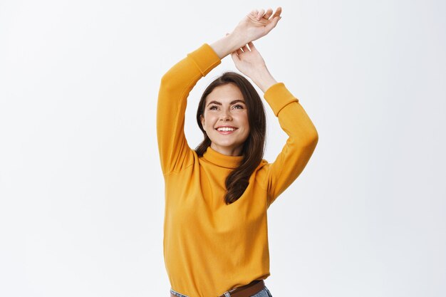 Sonriente mujer feliz levantando las manos sueltas con emociones despreocupadas y relajadas, de pie contra la pared blanca en ropa casual