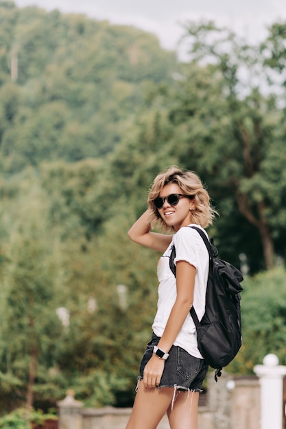 Sonriente mujer feliz con peinado corto vestido con camiseta blanca y pantalones cortos con gafas de sol negras viajando en las montañas, buen día soleado, senderismo en las montañas